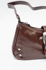 Bolso de hombro tipo baguette en color negro con acabado brillante. Presenta un diseño estructurado con una hebilla decorativa metálica en el frente y detalles de tachas plateadas en los laterales.