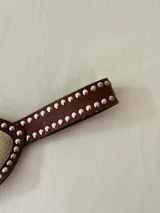 Cinto faja de cuero marrón con tachas metálicas plateadas en los bordes. Presenta una pieza central metálica con textura martillada y un dije en forma de gota con piedra natural.