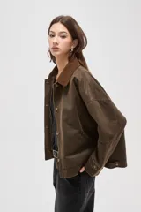 Campera de estilo encerado color marrón, con cuello de pana en tono contrastante, cierre frontal con cremallera y botones a presión, y bolsillos laterales. Presenta un diseño holgado con forro interior a cuadros.