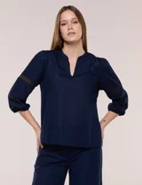 Blusa blanca de calce suelto y caída ligera, con escote en V y cuello Mao pequeño. Presenta mangas tres cuartos abullonadas con elástico en los puños y detalles de calado o puntilla encastrada. Espalda lisa con canesú y leve frunce.