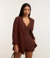 Blazer de sastrería color marrón, confeccionado en crepé, con diseño oversized y corte alargado. Presenta solapas clásicas, bolsillos frontales y detalle de pliegues en los puños.