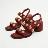 Sandalia de tacón bloque, confeccionada en gamuza color bordó. Presenta un diseño de tiras múltiples sobre el empeine, decoradas con apliques metálicos circulares en tono dorado. Cuenta con puntera cuadrada y cierre de hebilla ajustable en el tobillo.