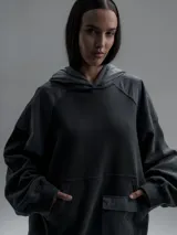 Hoodie oversize de color gris oscuro, confeccionado en frisa de algodón con recortes en textiles técnicos. Presenta capucha forrada, bolsillo frontal tipo canguro y un bolsillo adicional con solapa en la parte inferior.