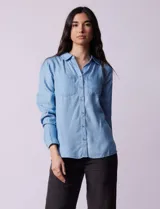 Camisa celeste de lyocell con cuello clásico, escote en V y cierre frontal con botones. Tiene dos bolsillos de parche en el pecho y mangas largas regulables con presilla y botón.