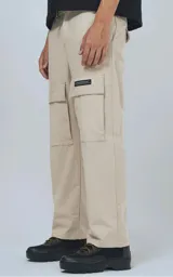 Pantalón cargo de corte regular confeccionado en tela ripstop color beige. Presenta siete bolsillos, incluyendo bolsillos laterales con solapa y etiqueta decorativa, y presillas en la cintura para ajuste.