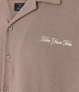 Camisa de manga corta, color marrón topo, con cuello camisero y corte amplio (boxy). Presenta bordados minimalistas en blanco en el frente, simulando un paisaje.