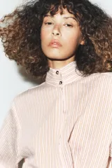Camisa de corte oversize con estampado de rayas verticales, cuello subido y cierre frontal con botones. Presenta mangas largas con puños y un diseño cruzado en la parte delantera.