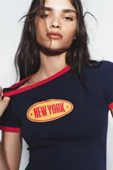 Camiseta de cuello redondo y manga corta, color azul marino con ribetes en color rojo en cuello y mangas. Presenta un estampado frontal ovalado con el texto New York en color naranja.