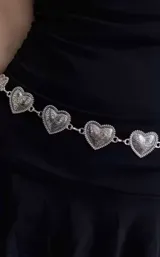 Cinto de cadena metálica plateada compuesto por eslabones en forma de corazón con detalles grabados en relieve.
