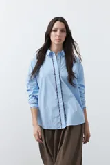 Camisa blanca de manga larga con detalles bordados en contraste a lo largo de la botonadura, el cuello y los puños. Presenta una silueta relajada y un diseño clásico con un toque artesanal.