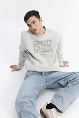 Buzo de cuello redondo color beige, con estampa frontal tipográfica de la marca Mistral en tonos grises. Presenta un diseño de calce regular con puños y ruedo acanalados.