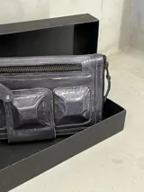 Billetera tipo clutch color gris oscuro de cuero con cierre superior y bolsillos frontales con solapa.