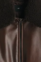 Campera de estilo aviador confeccionada en tejido revestido con acabado de cuero sintético. Presenta cuello de sherpa en contraste, cierre frontal de cremallera, hombros caídos y bolsillos delanteros de parche con solapa y botón a presión. Incluye elástico fruncido en el bajo y puños ajustables con botón a presión.