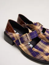 Zapatos tipo guillermina con punta cuadrada, confeccionados en lona resistente con estampado de cuadros escoceses en tonos beige, rojo y negro. Presentan una correa ajustable con hebilla metálica color bronce y una suela con taco de 4 cm.