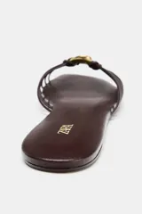 Sandalia plana de mujer, color marrón oscuro, con puntera cuadrada. Presenta un diseño de tiras finas que se unen en el centro con un adorno anillado metálico dorado.