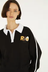 Buzo negro de felpa con cuello polo y solapa blanca, mangas largas con franjas blancas en los laterales y estampado en el pecho con la palabra "RIO" y detalles gráficos.