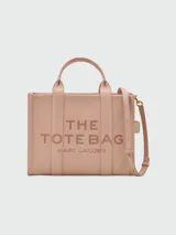 Cartera tipo tote de cuero granulado color tostado, con diseño estructurado y rectangular. Presenta doble asa superior fija y una correa larga ajustable y extraíble. En el frente, destaca el logo estampado con la inscripción 'THE TOTE BAG MARC JACOBS'.
