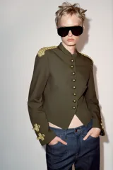 Cazadora militar de color verde oscuro, de cuello subido y manga larga. Cuenta con detalles de alamares y cordones dorados en los hombros y puños, y cierre frontal con botones dorados metálicos.