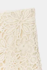 Falda midi de color crudo, con diseño combinado que presenta una parte superior de crochet con motivos florales y una parte inferior de tela fluida con textura arrugada. Incluye forro interior y cierre lateral con cremallera oculta.