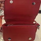 Cartera tipo bandolera de tamaño pequeño, color rojo, con solapa frontal y letras doradas estampadas que dicen 'THE MINI BAG'. Incluye una correa de tela ajustable y herrajes metálicos.