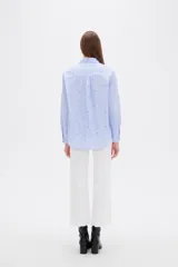 Blusa de corte holgado con cuello camisero y cierre frontal de botones. Presenta un diseño de rayas verticales finas en color azul y blanco, decorado con pequeños bordados dispersos.