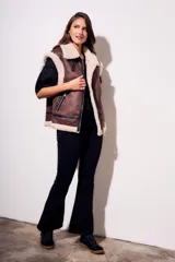 Chaleco oversize estilo gamulán en color marrón, confeccionado en cuero vegano con forro y solapas de corderito sintético en contraste. Posee cierre frontal metálico, bolsillos con cremallera y detalles de correas con hebillas en el cuello.