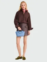 Cartera tipo tote de tamaño extra pequeño, confeccionada en cuero granulado color celeste. Presenta una estructura rígida con base trapezoidal, doble asa corta fija y cierre de solapa con botón a presión metálico dorado con logo grabado.