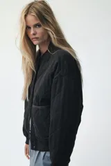 Campera bomber negra, acolchada con patrón de rombos. Presenta cuello, puños y bajo acanalados. Tiene cierre frontal con botones a presión y dos bolsillos de parche en la parte delantera.