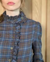 Camisa de manga larga con estampado de cuadros en tonos marrones y azules. Presenta cuello alto con volados, cierre frontal con botones y puños con volados.