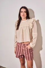 Blusa de color arena con diseño romántico, presenta volados en los hombros, detalles de encaje en el pecho y mangas largas con puños elásticos.