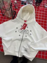 Buzo con cierre frontal, color crema, con diseño estampado de estrellas y logo en el pecho. Cuenta con capucha, bolsillos tipo canguro y tirador de cierre en forma de estrella.