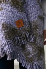 Ruana tejida de punto con diseño teñido artesanalmente en tonos lila y marrón, con terminación de flecos en el ruedo y etiqueta de cuero sintético en el frente.