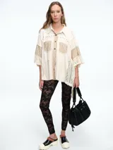 Camisa de corte oversize en color crudo, confeccionada con una mezcla de texturas que incluye paneles de encaje, detalles bordados y aplicaciones de tachas metálicas. Presenta mangas amplias tipo murciélago, cuello clásico y cierre frontal con botones.