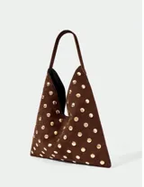 Bolso de hombro con diseño triangular, confeccionado en gamuza marrón y decorado con tachas metálicas doradas circulares en toda su superficie.