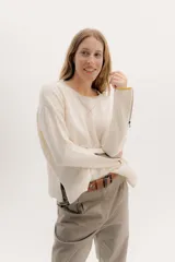 Buzo de lambswool color beige, con escote redondo y detalle en V. Presenta cierres metálicos en los puños que revelan un interior en contraste de color marrón y ribete rojo. Tiene tajos laterales para mayor comodidad y un fit amplio.