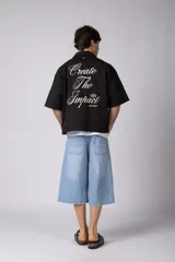 Camisa de manga corta negra, de corte boxy, con cuello camisero, cierre frontal de botones y bordado circular en el pecho.