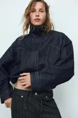 Cazadora de denim color azul oscuro, con cuello solapa, mangas largas tipo murciélago y bolsillos de vivo en el delantero. Presenta un bajo elástico ajustable con cordones y cierre frontal con cremallera y botones a presión ocultos.