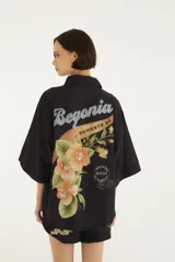 Camisa de manga corta negra con cuello camisero, cierre frontal de botones y diseño holgado, decorada con estampados botánicos y gráficos de estilo vintage.