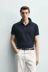 Remera polo de punto canalé color azul marino, de corte regular fit, con cuello solapa y cierre frontal de botones a presión. Cuenta con mangas cortas con ribetes blancos en los bordes del cuello y de las mangas.