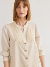 Camisa de corte recto y manga larga, confeccionada en algodón blanco. Presenta un canesú superior con bordado inglés floral y detalle de placa metálica con logo en la espalda.