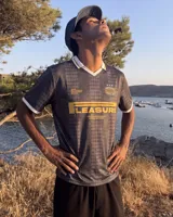 Camiseta de fútbol estilo retro de manga corta, color negro con diseño texturizado a rayas verticales, cuello en V blanco con detalle en mostaza y bordes de mangas a juego. Presenta estampados gráficos en el frente con la inscripción "PLEASURE" y emblemas deportivos.