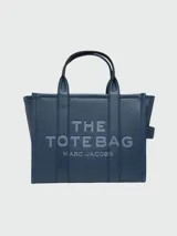 Cartera tipo tote de cuero granulado color tostado, con diseño estructurado y rectangular. Presenta doble asa superior fija y una correa larga ajustable y extraíble. En el frente, destaca el logo estampado con la inscripción 'THE TOTE BAG MARC JACOBS'.