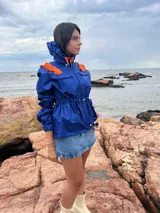 Piloto impermeable color azul eléctrico con detalles naranjas en los hombros, capucha integrada, cordón regulable en la cintura y mangas con volumen y puños elastizados.