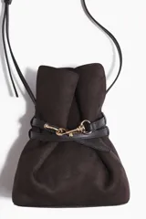 Cartera tipo bombonera de color marrón oscuro, confeccionada en tejido revestido con acabado mate. Presenta un diseño fruncido en la parte superior ajustado por una tira delgada con cierre de mosquetón metálico dorado. Incluye una correa larga y fina ajustable para llevar cruzada.