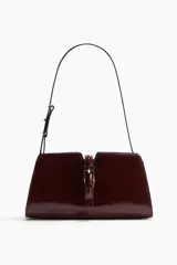 Cartera bandolera bordó con acabado charol, diseño rectangular y solapa frontal decorada con una tira y hebilla metálica dorada. Cuenta con una correa fina ajustable para llevar al hombro o cruzada.