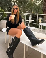 Botas de caña alta confeccionadas en ecocuero negro, con diseño de tres hebillas metálicas laterales y suela plana.