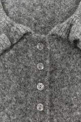 Chaqueta de punto gris con cuello solapa, manga larga y cierre frontal con botones. Presenta detalles de volantes en el cuello, los puños y el ruedo.