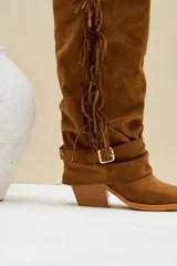 Botas altas estilo texana de gamuza color camel, con taco cuadrado y puntera fina. Presentan flecos largos colgando a lo largo del lateral de la caña y una correa con hebilla metálica y argollas alrededor del tobillo.