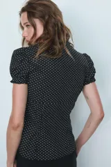 Camisa negra con estampado de lunares blancos, cuello redondo con escote en pico, mangas cortas abullonadas y detalle de volantes frontales. Cierre frontal con botones.