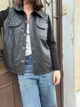 Campera negra con cuerpo de cuero y mangas de lana. Tiene corte recto, cuello camisero, cierre frontal con botones dorados, dos bolsillos con solapa en el pecho y detalles de tachas doradas.